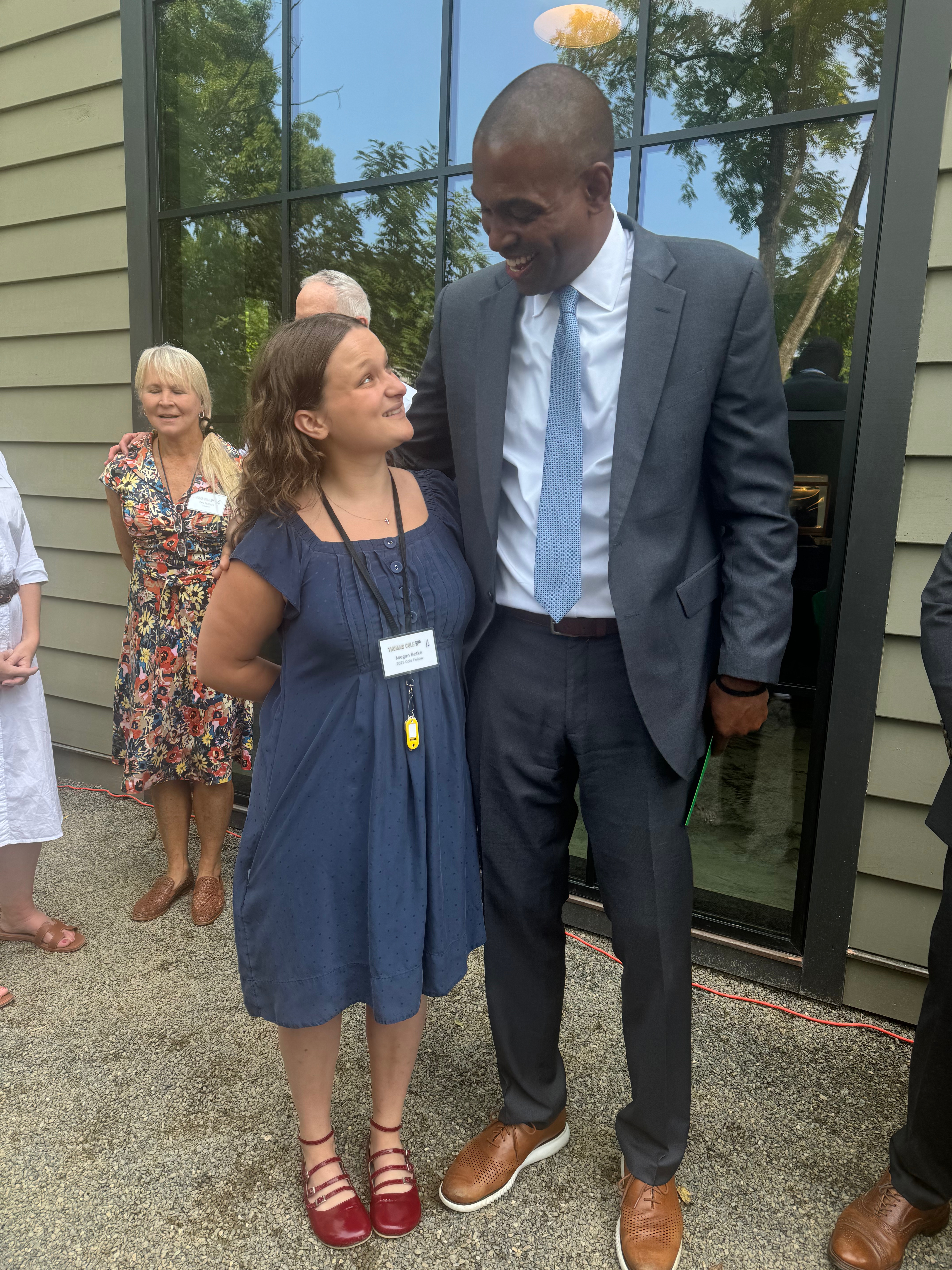 A woman in a blue dress with red shoes stands next to a tall man in a suit, both smiling and engaged in conversation. In the background, other individuals are partially visible, with trees and a large window providing a natural backdrop.